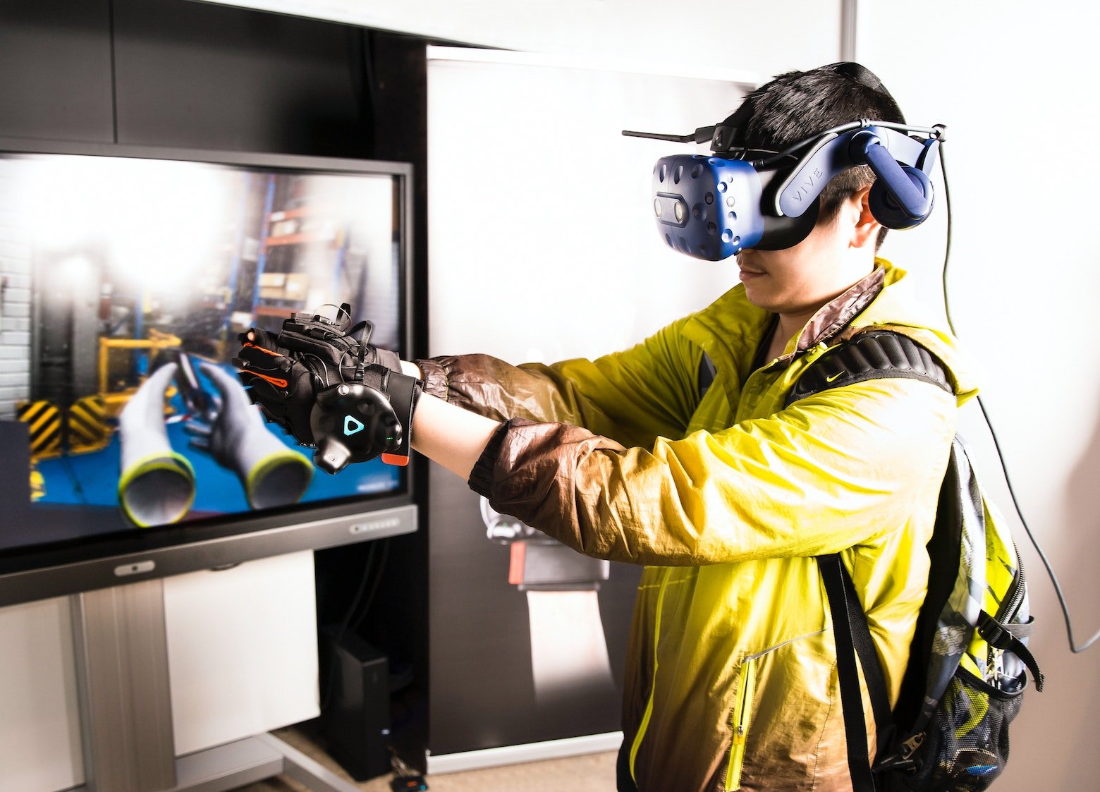 A man wearing a headset and VR gloves. A screen in front of him depicts virtual gloves that he is controlling.