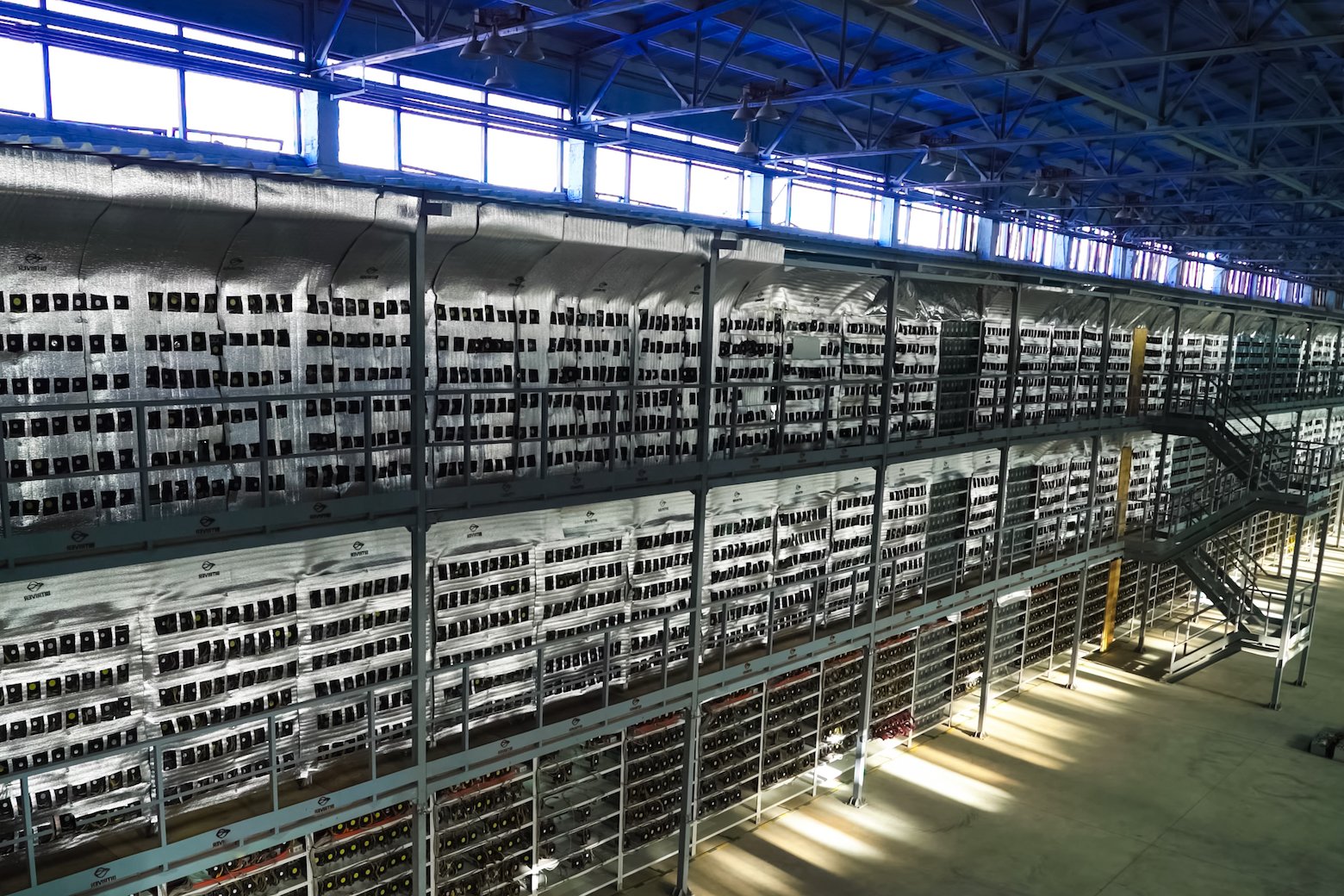 The inside of a large warehouse showing thousands of cryptocurrency miners stacked in large metal racks three levels high.
