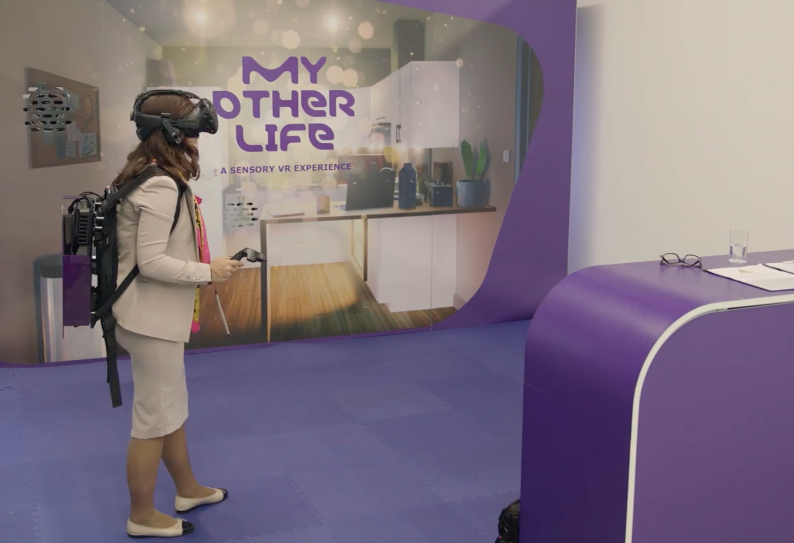 A woman wears a headset and holds a controller in front of a backdrop that depicts a kitchen. Text overlaid on the backdrop reads, “My Other Life: A Sensory VR Experience.”