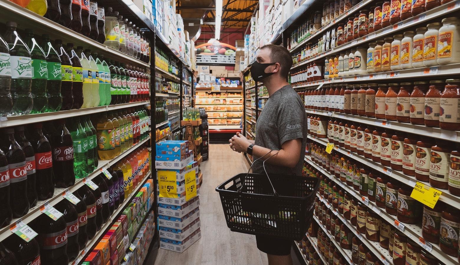 A young man wearing a cloth mask and a gray shirt stands in a grocery store with a shopping basket on his forearm. He is looking at shelves full of soft drinks.