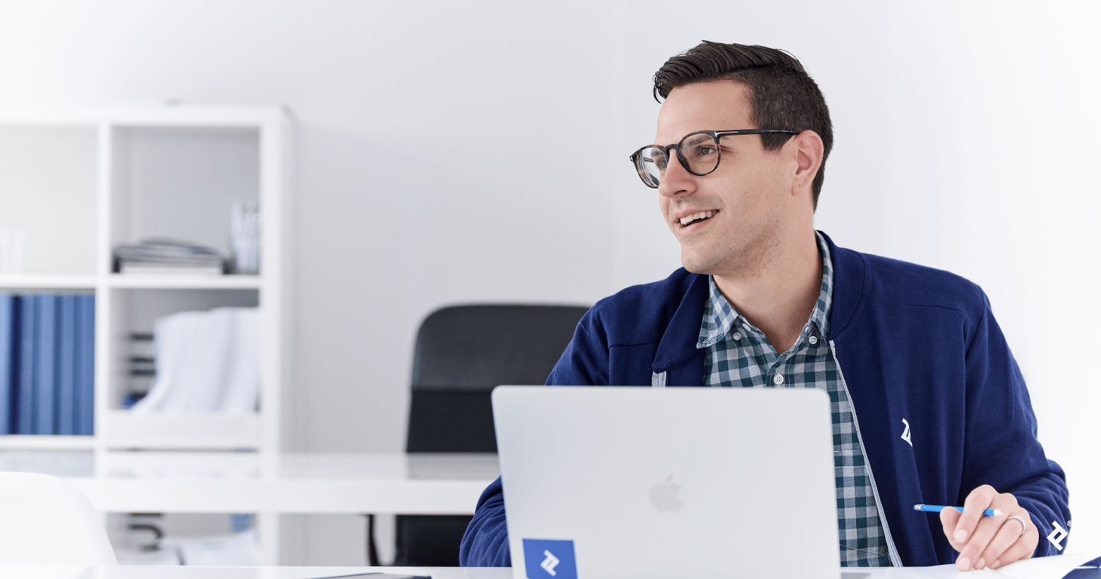 A man with dark hair and glasses wearing a Toptal-branded jacket sitting behind a laptop in a white office area.