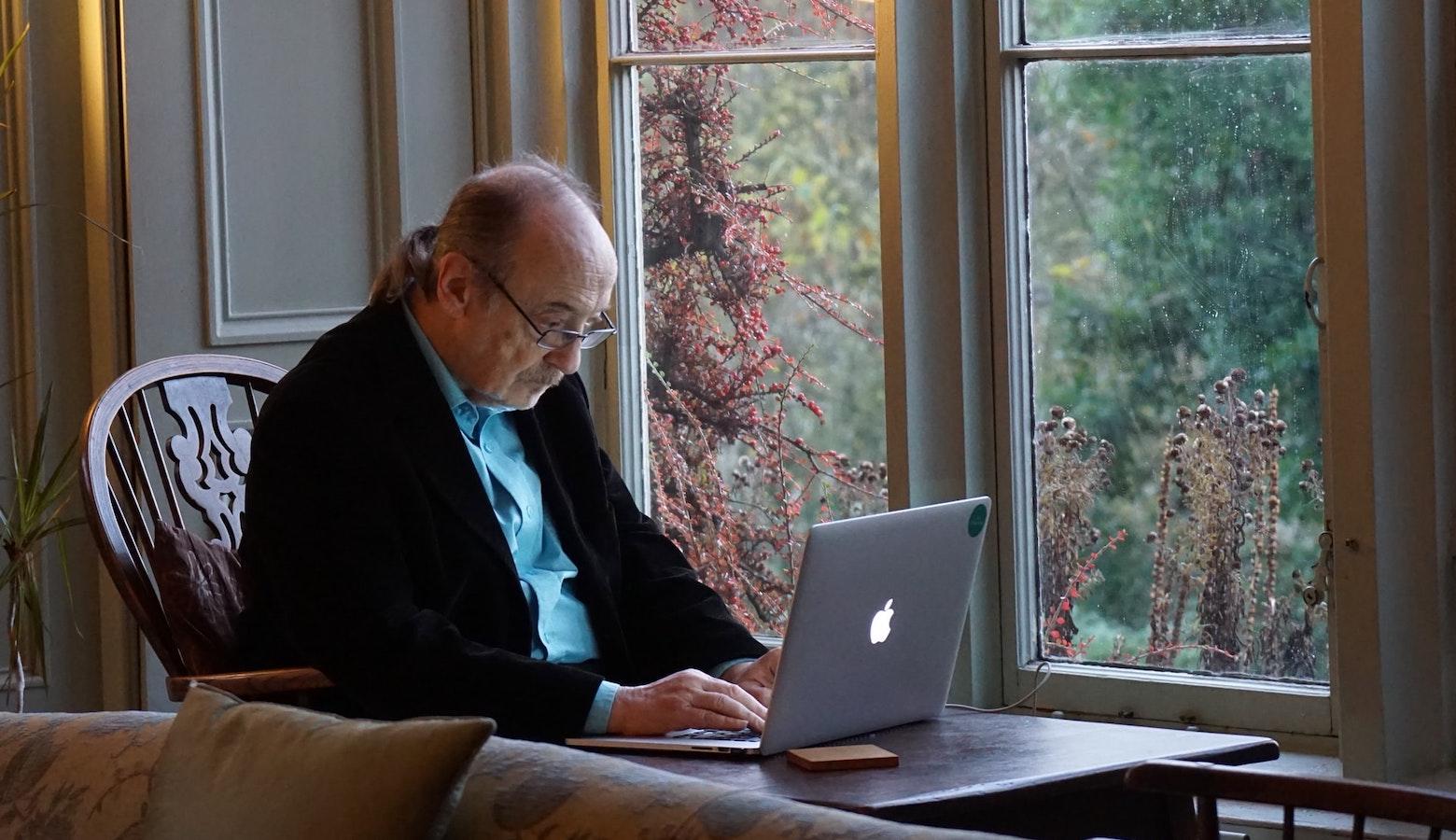 An older man sitting in a wooden chair at a table in front of a window. He is typing on an Apple laptop.