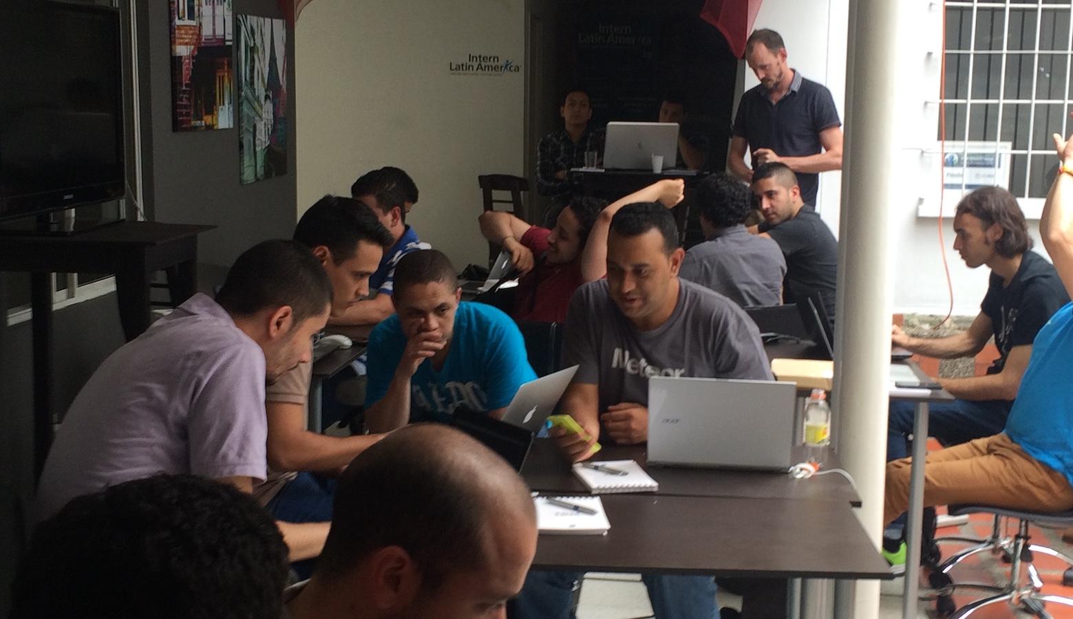 Approximately a dozen young people working with laptops at tables in small teams in an outdoor classroom. Two older men are in the back observing.