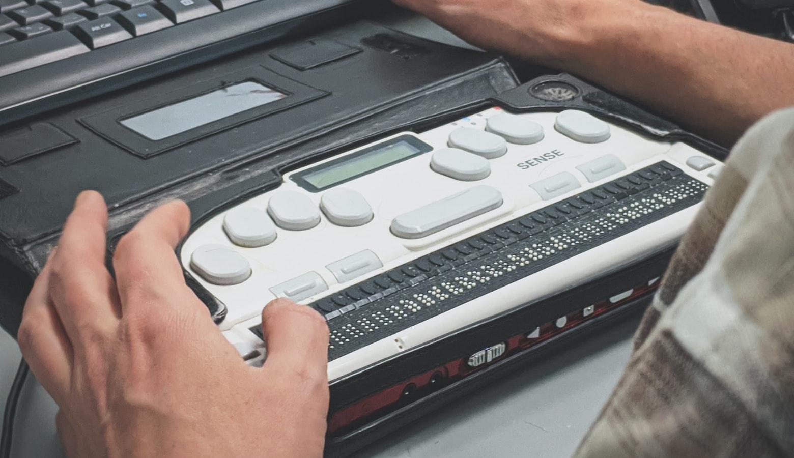 Close-up photo of a refreshable Braille display and Braille keyboard docked to a standard keyboard. A man is touching the Braille with his thumb.
