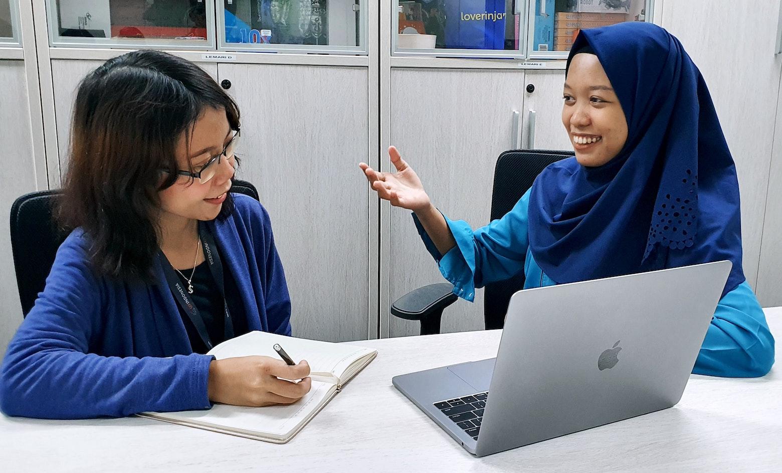Two women sit at a table turned toward each other with a laptop between them. The woman on the left, wearing a blue cardigan and glasses, is writing in a notebook as she conducts user research. The woman on the right, who is wearing a blue hijab, is speaking and gesturing as if to explain how she does something with the computer.