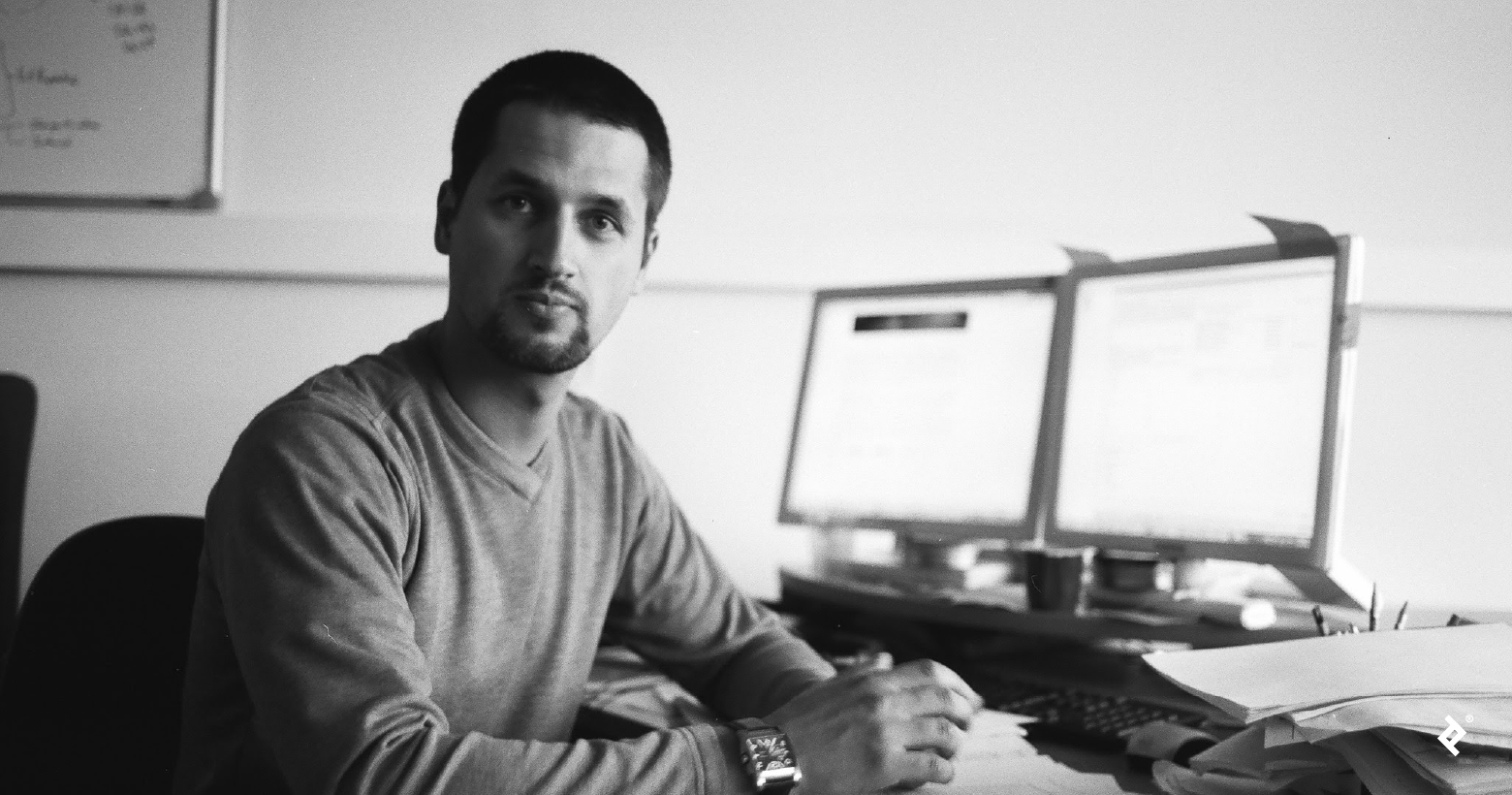 A black-and-white photo of a man with short hair and a goatee, wearing a long-sleeved T-shirt and looking at the camera. He is sitting at a desk with computer monitors behind him.