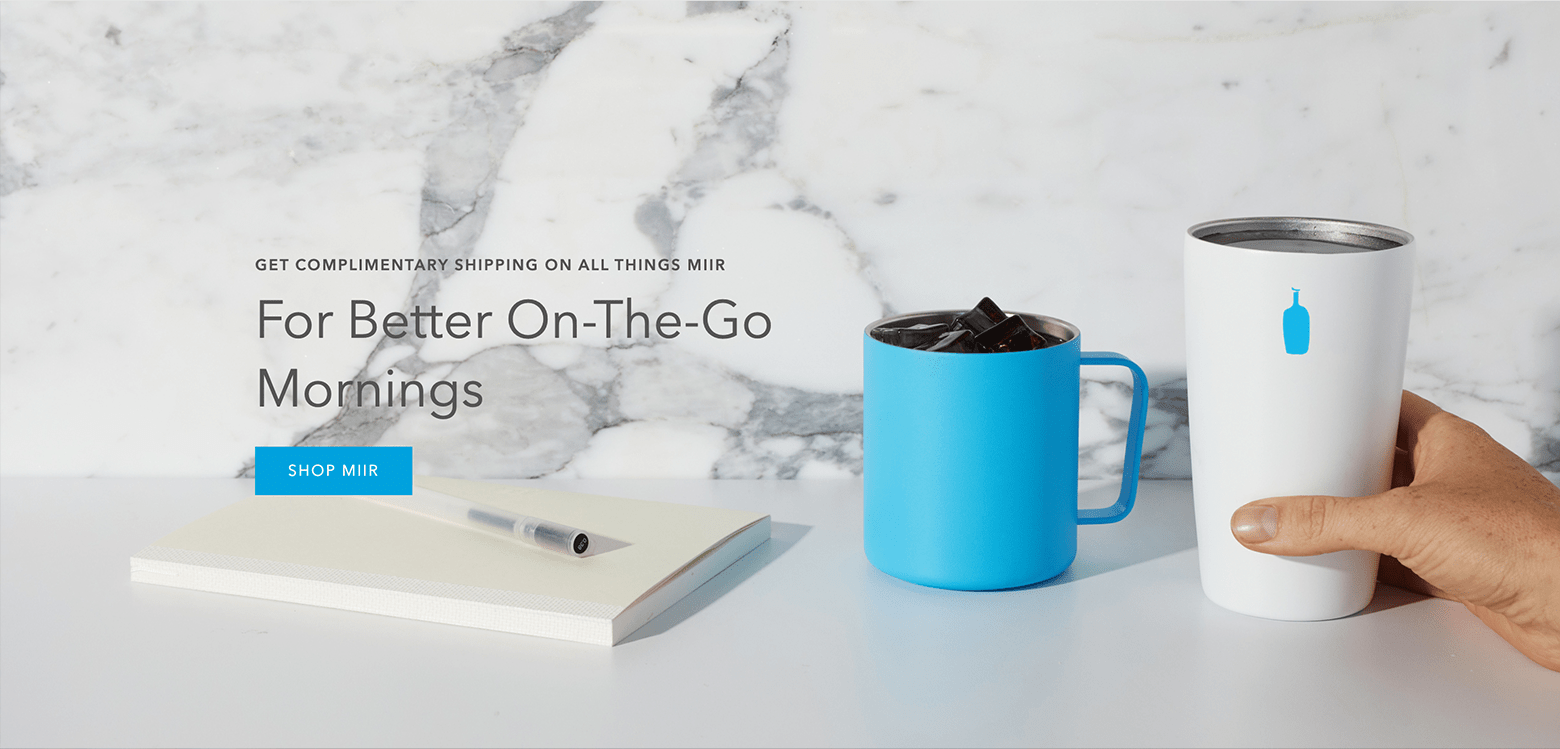 An image of a notebook and pen, a blue mug, and a white mug with a blue logo on a white table. A hand holds the white mug, and the image has a marble backdrop. Text overlaid on the image reads, "Get complimentary shipping on all things MiiR." Larger text below this reads, "For Better On-The-Go Mornings," above a blue "Shop MiiR" button.